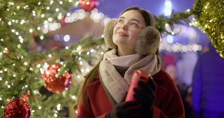A young beautiful girl in a red coat walks in the evening at the European Christmas Market and holds a thermos cup, standing against the backdrop of a Christmas tree with garlands and illuminations.