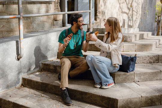 Man and woman clinking cups and laughing while holding croissants and sitting on stairs