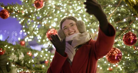 A young beautiful girl in a red coat walks in the evening at the European Christmas Market making a video call standing against the backdrop of a Christmas tree with garlands and illuminations