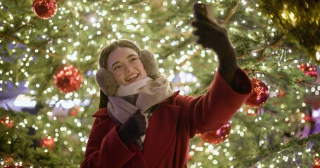 A young beautiful girl in a red coat walks in the evening at the European Christmas Market making a video call standing against the backdrop of a Christmas tree with garlands and illuminations