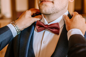 Man in formal attire adjusts his red bow tie with assistance, showcasing a moment of preparation for a special event, emphasizing elegance and attention to detail
