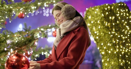 A young beautiful girl in a red coat stands near a Christmas tree with garlands and illuminations in the evening at the European Christmas Market and touches the Christmas tree decorations.