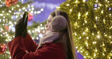 A young beautiful girl in a red coat walks in the evening at the European Christmas Market and takes pictures on a phone while standing against Christmas tree with garlands and illuminations.