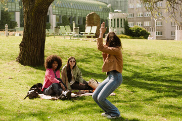 Two female friends are smiling and sitting on a blanket while looking at a female friend who is showing while standing opposite them on the grass