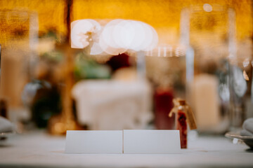 Elegant table setting with blurred background, featuring two blank name cards on a white tablecloth, surrounded by decorative glassware and warm ambient lighting for special occasions