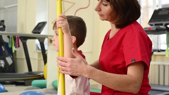 A physical therapist works with a child on posture exercises. A professional osteopath helps a boy do scoliosis exercises with gymnastic sticks.