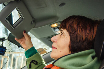 Mature woman sitting in car, lifting sun visor with mirror, closeup, careful preparation, driver safety, senior mobility