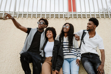 A group of four friends stand and lean on a wall while smiling and looking in the direction one of...