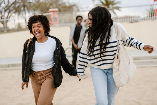 Two female friends holding hands and laughing as they walk with a male friend walking behind them