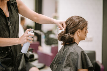 Female hairstylist applying hairspray to elegant updo hairstyle on client in modern salon, showcasing professional hairdressing techniques and stylish atmosphere