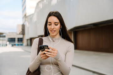 Female athlete typing on phone and listening to earphones while carrying a bag on her shoulder