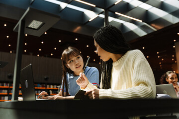 Female student listening to female student talking and holding a notebook while they hold pens and sit at a table in front of a laptop