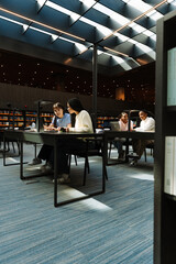 Female student talking to female student holding pen and notebook while they sit at table and look at laptop