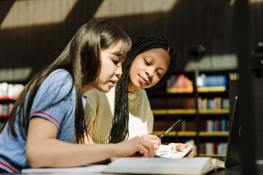 Female student holding a pen and listening to a female student talking while they are reading a book and sitting at a table
