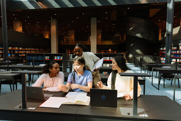 A group of three female students are sitting at a table and talking while another female student stands next to them and they look at a laptop