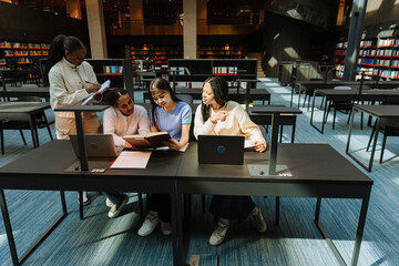 A group of three female students are reading a book and sitting at a table while another female student stands next to them and writes in a notebook