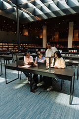 A female student stands next to a group of three female students sitting at a table while they read a notebook and a book and smile