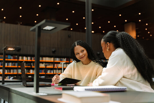Female student holding a pen and smiling while listening to a female student sitting next to her at the table