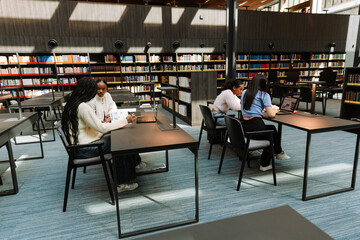 A group of four female students sit at desks in pairs while looking at a laptop and talking