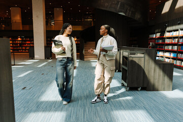 Female student holding notebooks and talking to female student walking next to her and holding...