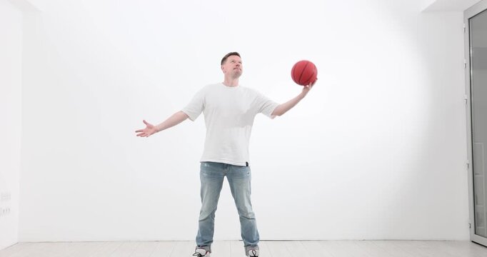 Man stands in spacious living room and passes ball between hands. Strong sportsman warms up muscles before starting training routine. Slow motion