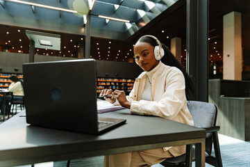 Female student looking at laptop and listening to headphones while sitting at desk and holding pen