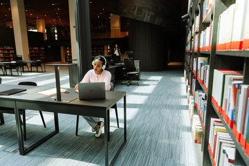 Female student holding a pen and listening to headphones while sitting at a desk in front of a laptop