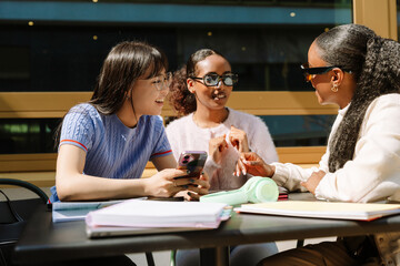 Two female students laughing and listening to a female student talking while they sit at a table and one of them is holding a phone