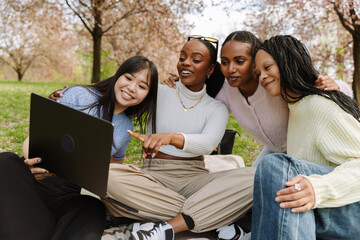 A group of four female students sit and laugh while hugging and looking at a laptop, one of them pointing at it while the other shows it to them