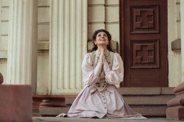 Fototapeta premium An elegant woman in a vintage dress sits next to an old stone column with a very happy expression on her face.