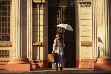 Fototapeta premium A woman in an antique dress with an umbrella and a suitcase stands in front of the doors of an old cathedral.