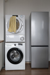Stacked washing machine and dryer beside a tall grey refrigerator in a laundry room with a yellow towel hanging on the dryer