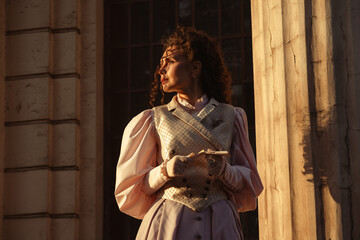 Fototapeta premium Portrait of a woman in vintage clothing, illuminated by warm sunlight, against a backdrop of ancient architecture.
