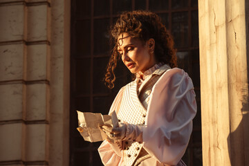 Fototapeta premium A woman in a vintage dress reads a letter near the columns of an ancient cathedral.