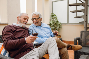 Obraz premium A man smiles and talks to his male friend who listens while they hold glasses and sit on a chair