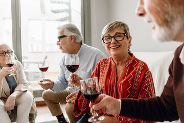 A woman smiles while sitting on a sofa next to two male friends and a female friend sitting on a chair while they hold glasses and talk