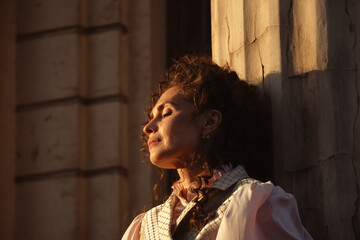 Fototapeta premium Portrait of a woman in vintage clothing, illuminated by warm sunlight, against a backdrop of ancient architecture.