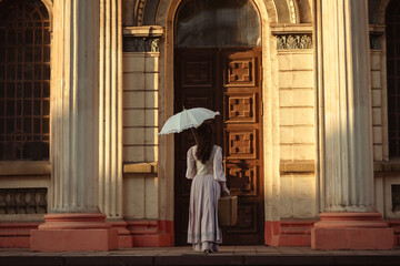 Fototapeta premium A woman in an antique dress with an umbrella and a suitcase stands in front of the doors of an old cathedral.