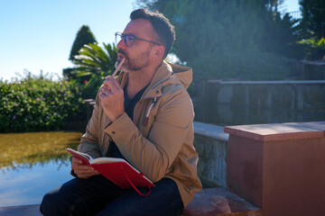 Man in glasses thinking outdoors, holding pencil, writing ideas in a red notebook by the water