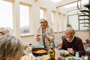A woman laughs and holds a spatula while standing at a table with a group of three friends sitting around it
