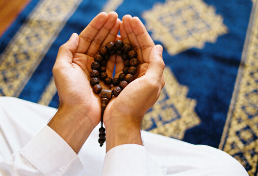 Person engaging in muslim pray while holding prayer beads, with an ornate mat on ground
