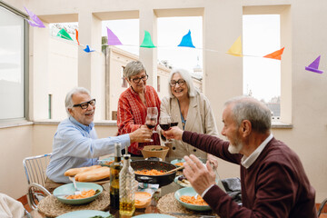 Two male friends sitting at a table clinking glasses with two female friends standing around while they laugh