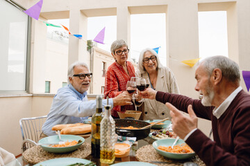 Two male friends are talking and sitting at a table while two female friends are standing next to them and smiling as they clink glasses