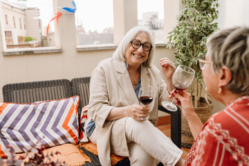 Woman sitting on a chair and laughing while listening to her female friend while they hold glasses