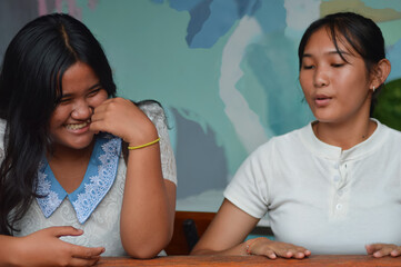 A close-up portrait of two teenage girls sitting at a wooden table, possibly in a garden cafe. One in a lace top laughs heartily covering her mouth while her friend in a henley talks.