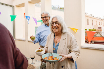 A woman carries plates and bowls while a man stands behind her at the table and they laugh