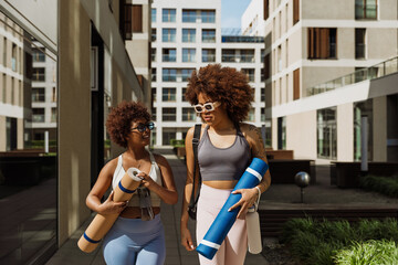 A woman smiles and talks to a female friend walking alongside her as they carry mats and bottles