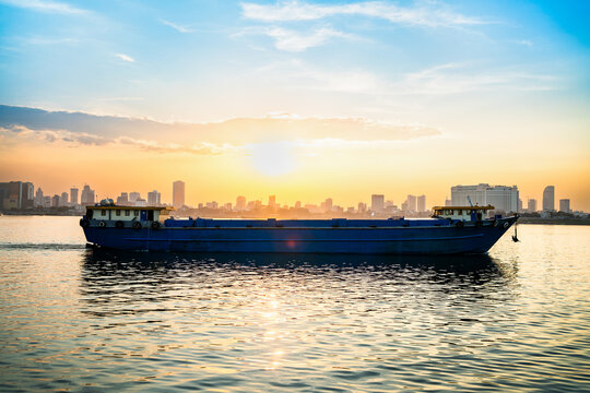 Cargo ship and dry bulk vessel at sunset. Export or import in Asia. Grain or wheat transportation. Phnom Penh city skyline in the background in Cambodia. Food or general carrier in Mekong river.