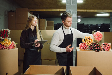 Two caucasian florists packing tulips in warehouse, filling cardboard boxes, checking colorful wrapped bouquets, using tablet for orders, warm cooperative mood under industrial lights