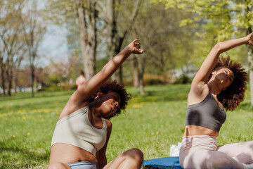 A woman sits next to a female friend who is sitting on a mat while they stretch their arms out to the side above their heads with their eyes closed